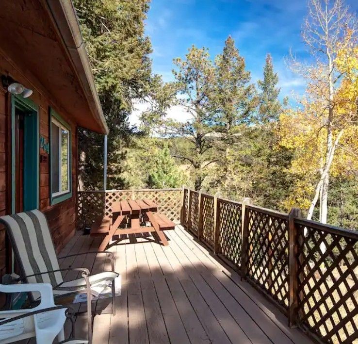 Wooden porch with picnic table and chairs overlooking a forested area under a bright blue sky.